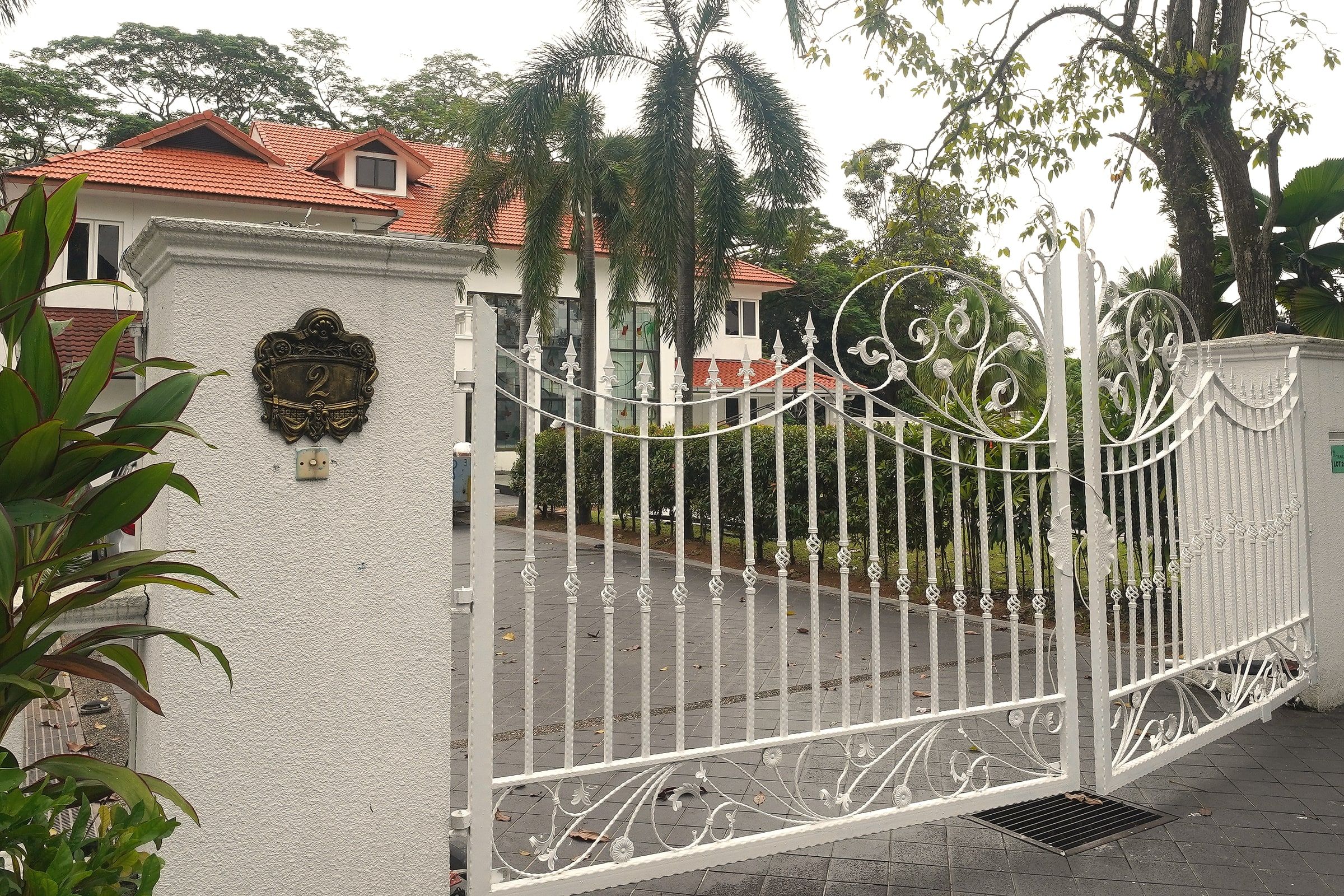 The open white gate and brass crest at Trivelle Living, with the red-roofed bungalow behind the hedges